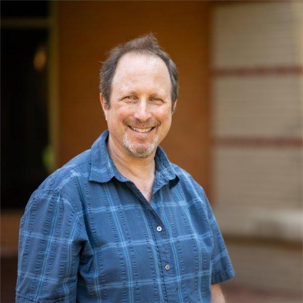 Joel Stillerman in blue short-sleeve shirt standing against brick wall